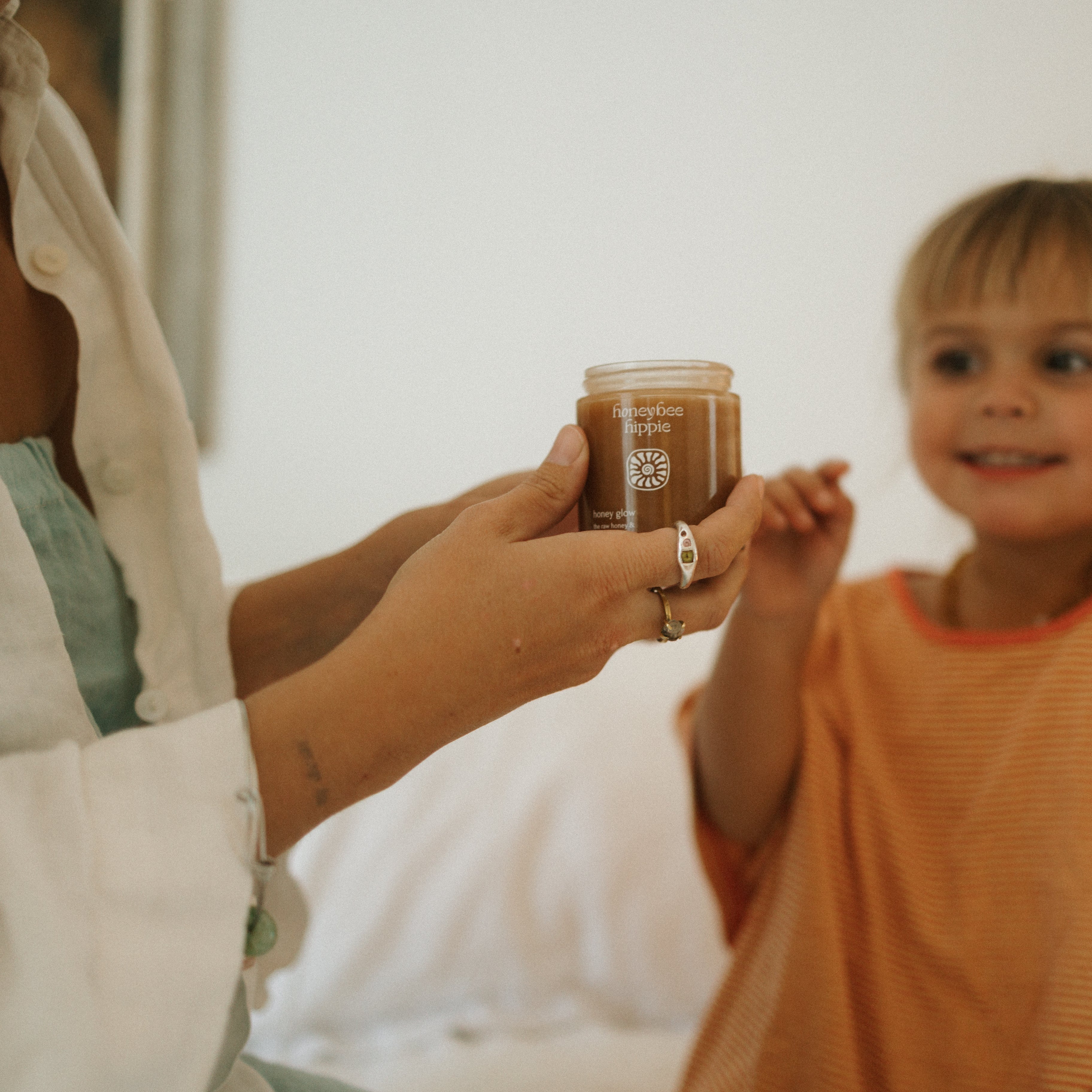 An adult hands a jar of Honeybee Hippie’s honey glow to a smiling child in an orange shirt, both indoors against a white background—celebrating natural glowing skin.