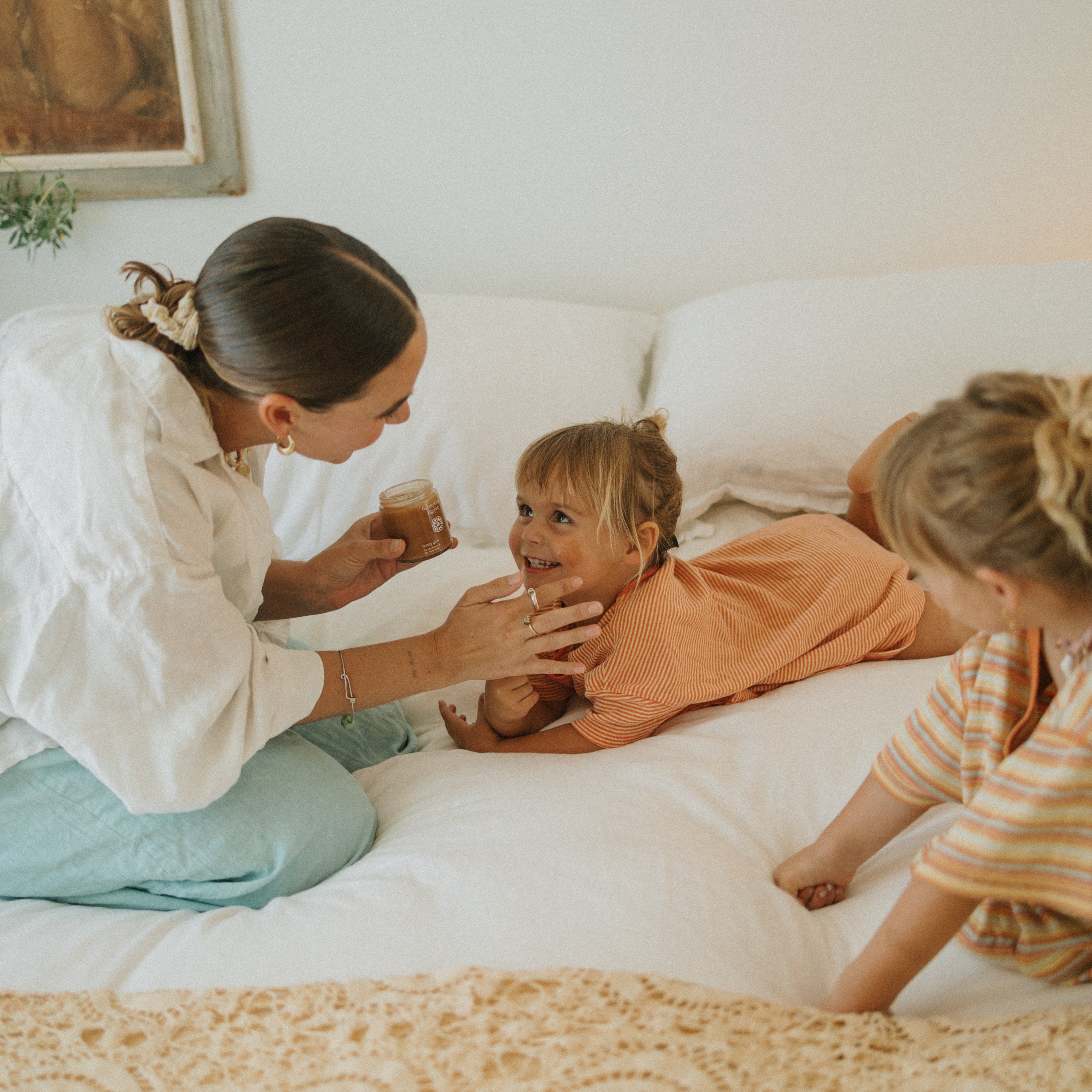 A woman applies Honeybee Hippie’s honey glow cream to a smiling young girl’s face as another child sits nearby, all enjoying a happy, playful moment in a cozy, sunlit room.