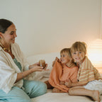 A woman with glowing skin sits on a bed, holding Honeybee Hippie's honey glow cleanser, smiling at two cheerful children in striped shirts beside her. A warm lamp glows softly in the background.