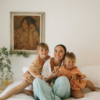 A woman with two girls in matching striped dresses sits on a bed, all showing natural glowing skin thanks to Honeybee Hippie's honey glow. A framed painting and a plant are visible in the background.