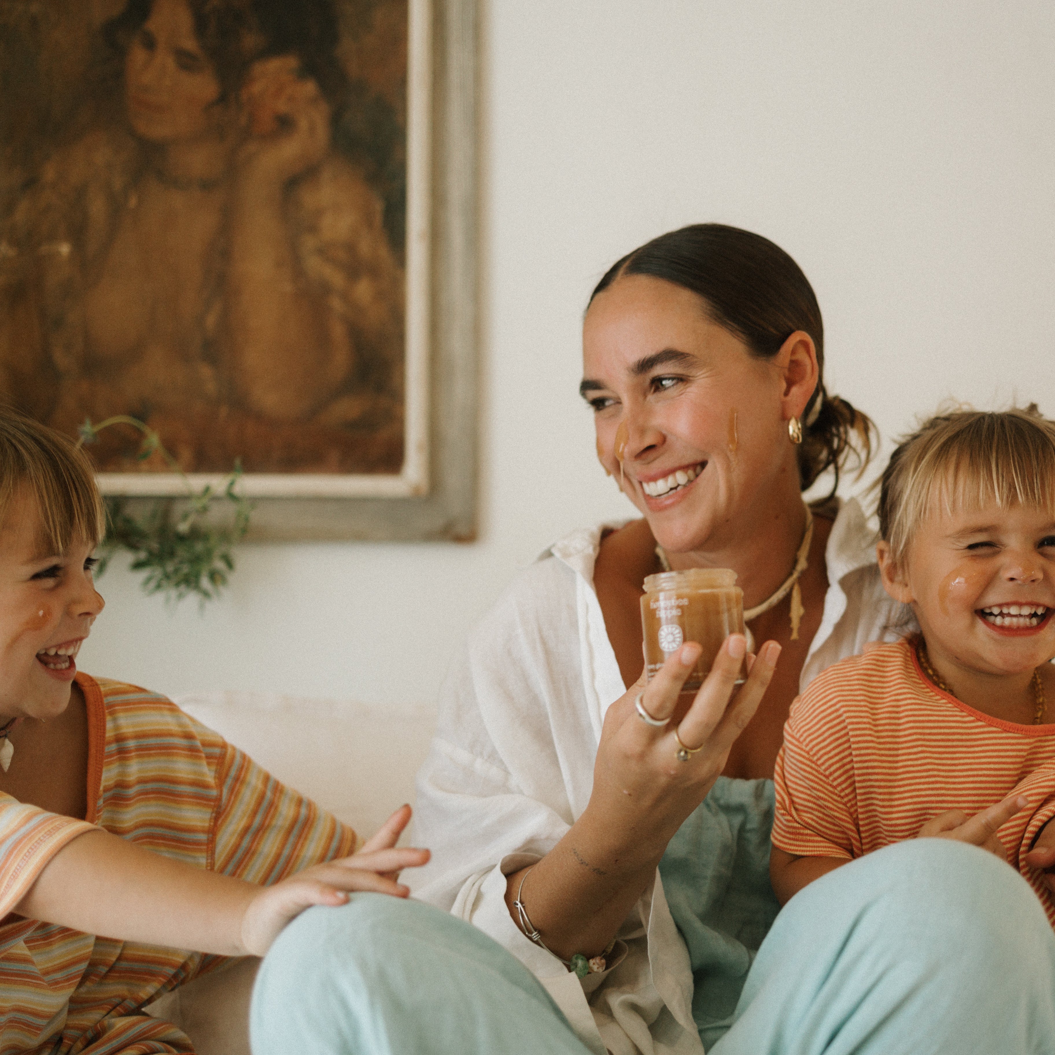 A woman smiles on a couch, holding a jar of Honeybee Hippie’s honey glow face cleanser as two laughing children in striped shirts sit beside her. A framed painting on the wall enhances their joyful moment.