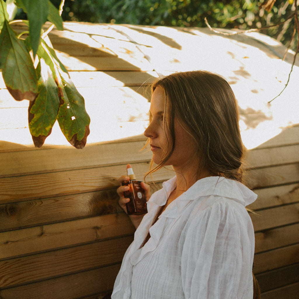 A woman in a white blouse stands by a wooden fence outdoors, holding a Honeybee Hippie rose hydrosol bottle as sunlight and leaves cast shadows—evoking the gentle promise of natural skin hydration.