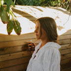 A woman in a white blouse stands by a wooden fence outdoors, holding a Honeybee Hippie rose hydrosol bottle as sunlight and leaves cast shadows—evoking the gentle promise of natural skin hydration.