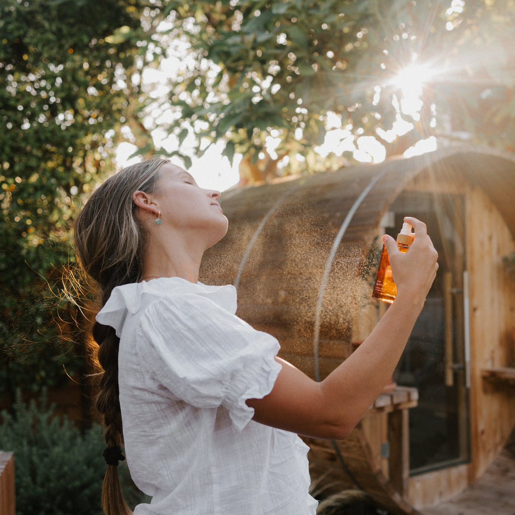 A woman with a long braid sprays Honeybee Hippie rose hydrosol onto her face outdoors near a wooden structure, sunlight streaming through the trees. She wears a white blouse, looking relaxed and refreshed.