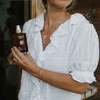 A woman in a white, ruffled blouse holds a brown bottle of Honeybee Hippie rose hydrosol near her chest, standing before a wooden wall. The scene evokes the fresh allure of floral mist. Her face is partially out of frame.