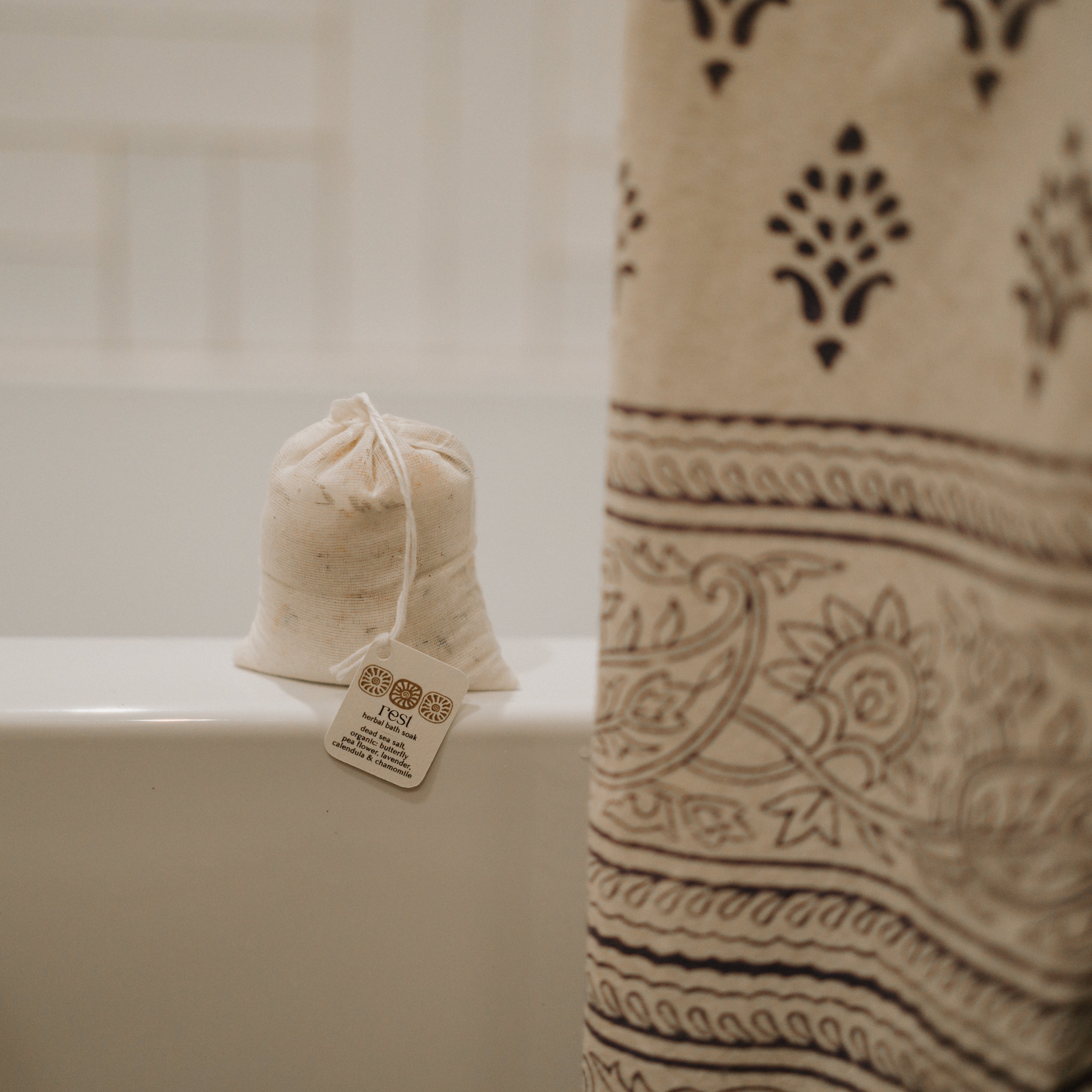 A small drawstring bag labeled "rest bath soak" by Honeybee Hippie sits on the edge of a white bathtub near a beige, intricately patterned shower curtain, with white tiled walls in the background.