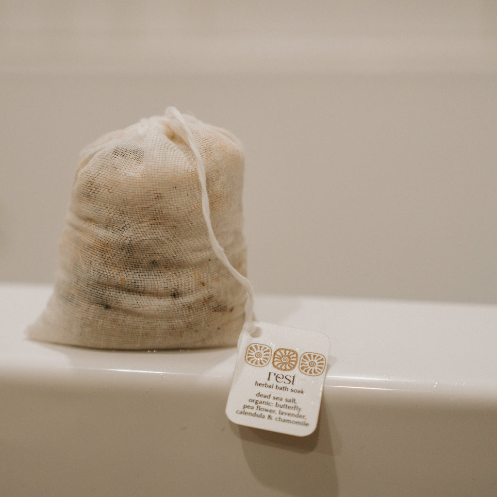 A mesh pouch of Honeybee Hippie’s rest bath soak sits on a tub’s edge. Its label lists calming ingredients like calendula, chamomile, and Dead Sea salt.