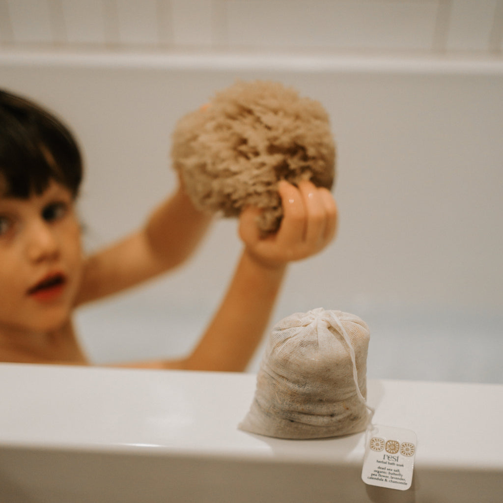A young child in the bathtub holds a large natural sponge, while Honeybee Hippie’s rest bath soak in a small muslin bag with label sits on the tub’s edge in the foreground.