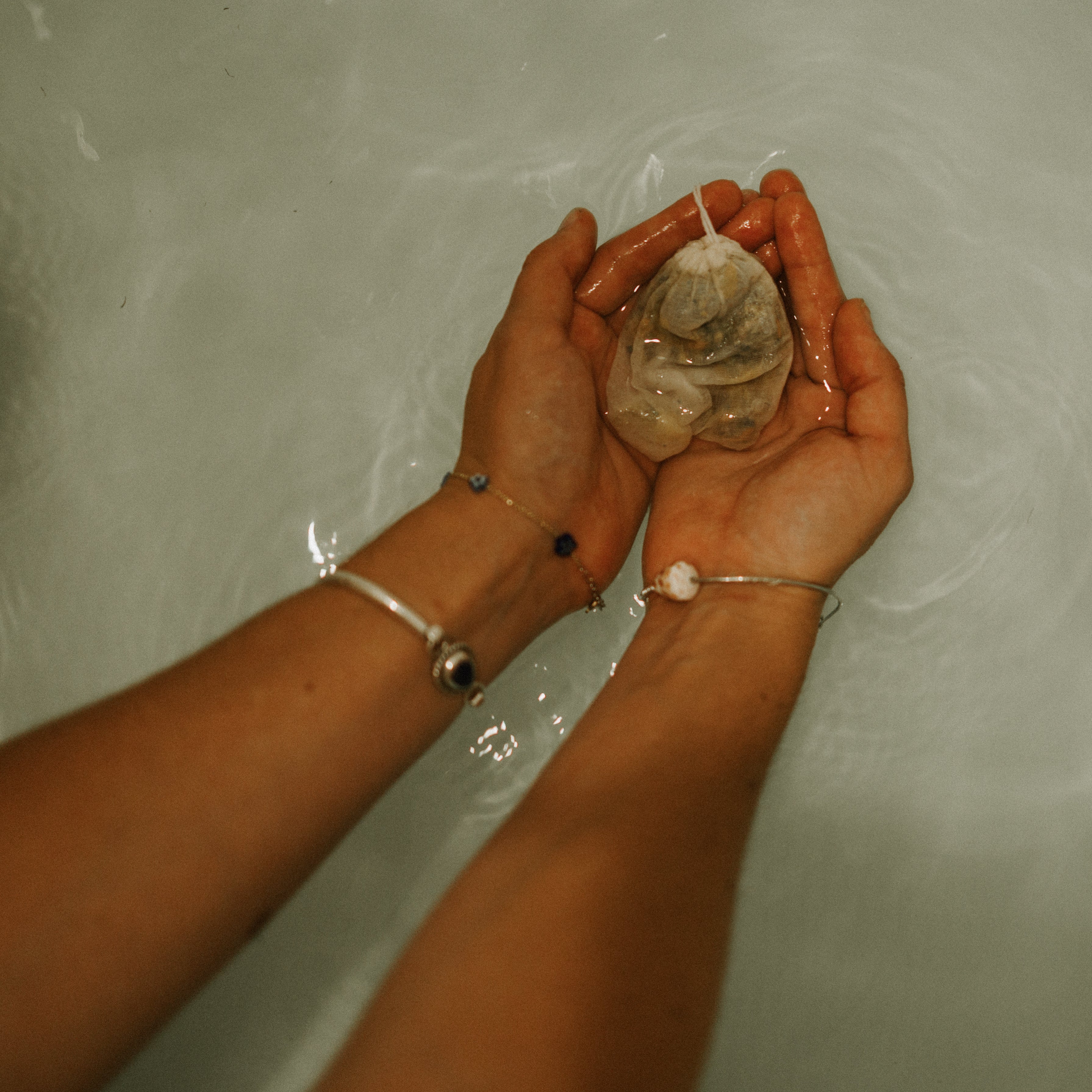 Two hands holding a Honeybee Hippie postpartum herbal sitz bath tea bag hover over rippling water, showcasing the organic blend for soothing relief.