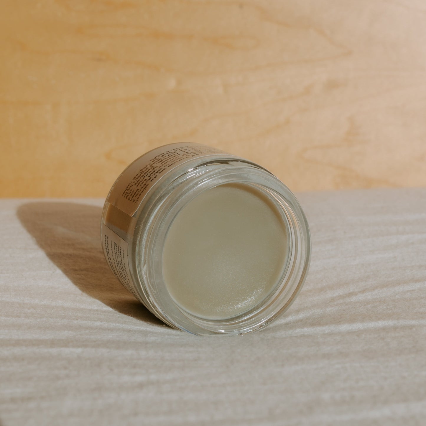 A glass jar of Honeybee Hippie's Miracle Mud, a tallow-based balm, lies on its side against a wooden background. The beige lid complements the light surface, and sunlight casts a gentle shadow, highlighting this natural skincare gem.