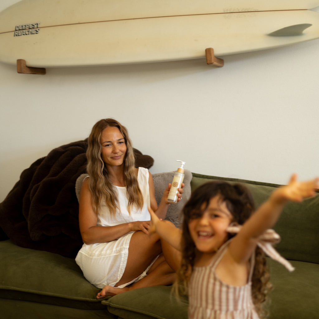 A woman in a white dress sits on a green couch holding a Honeybee Hippie magnesium butter jar, while a young child in striped clothes smiles and reaches toward the camera. A surfboard is visible on the wall behind them.
