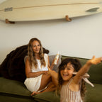 A woman in a white dress sits on a green couch holding a Honeybee Hippie magnesium butter jar, while a young child in striped clothes smiles and reaches toward the camera. A surfboard is visible on the wall behind them.