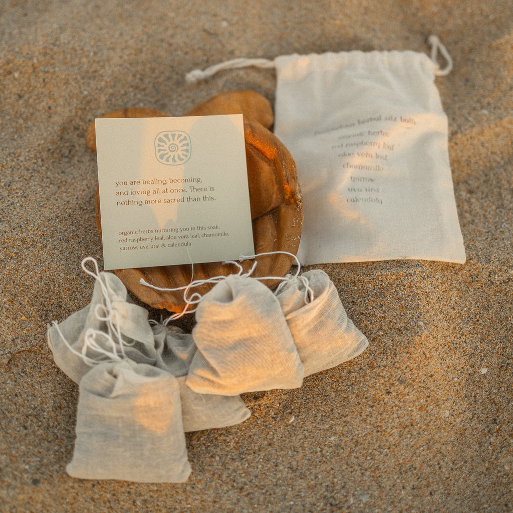Honeybee Hippie postpartum herbal sitz bath bags are nestled on sand beside a drawstring pouch and message card. A wooden bowl holds more of the organic herb bags, creating a calm, natural setup for soothing postpartum self-care.