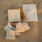 Honeybee Hippie postpartum herbal sitz bath bags are nestled on sand beside a drawstring pouch and message card. A wooden bowl holds more of the organic herb bags, creating a calm, natural setup for soothing postpartum self-care.