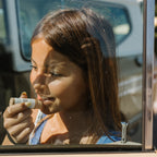 A young girl with long brown hair applies Honeybee Hippie vanilla & chamomile tallow lip butter while gazing out a car window, sunlight casting gentle shadows on her face.