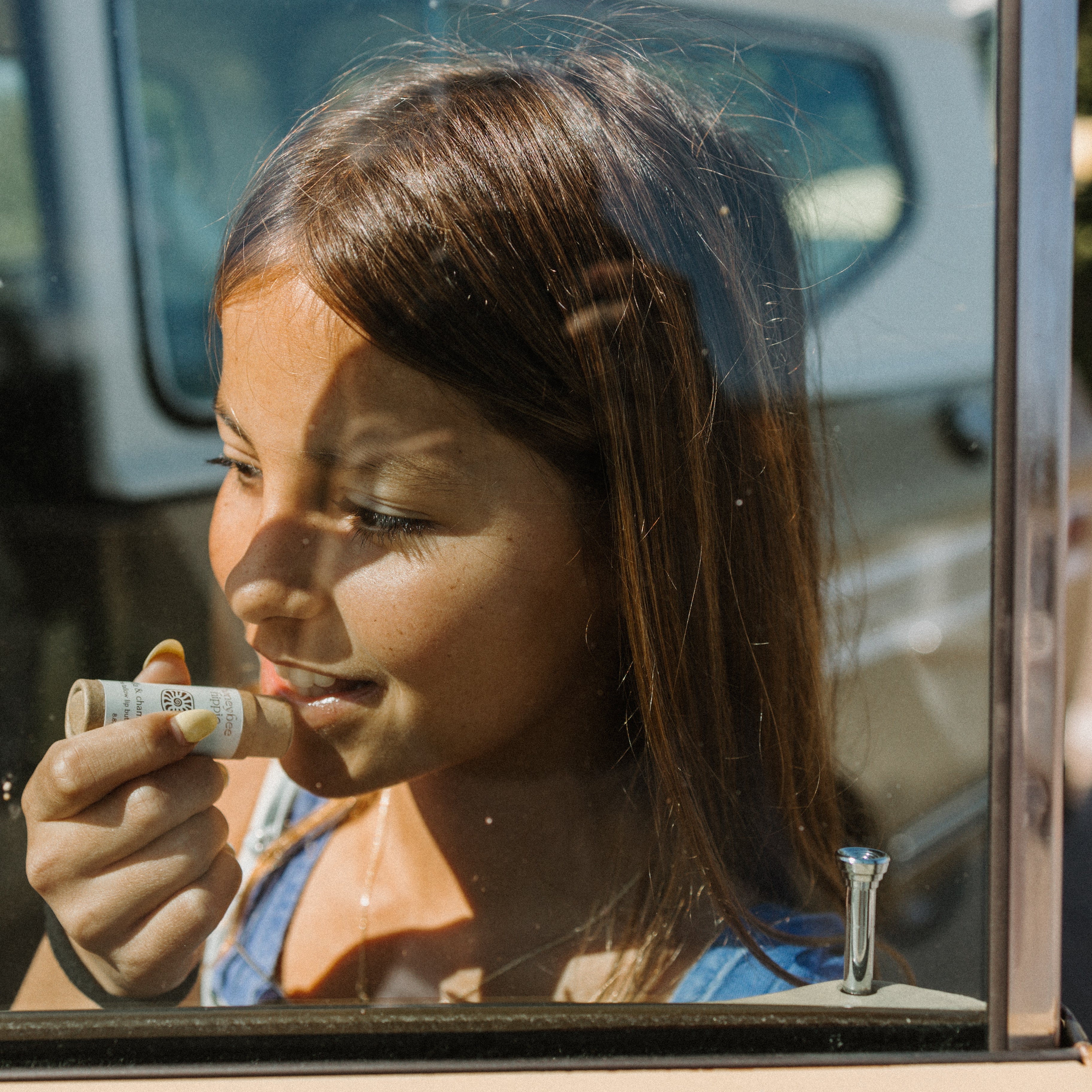 A young girl with long brown hair applies Honeybee Hippie vanilla & chamomile tallow lip butter while gazing out a car window, sunlight casting gentle shadows on her face.