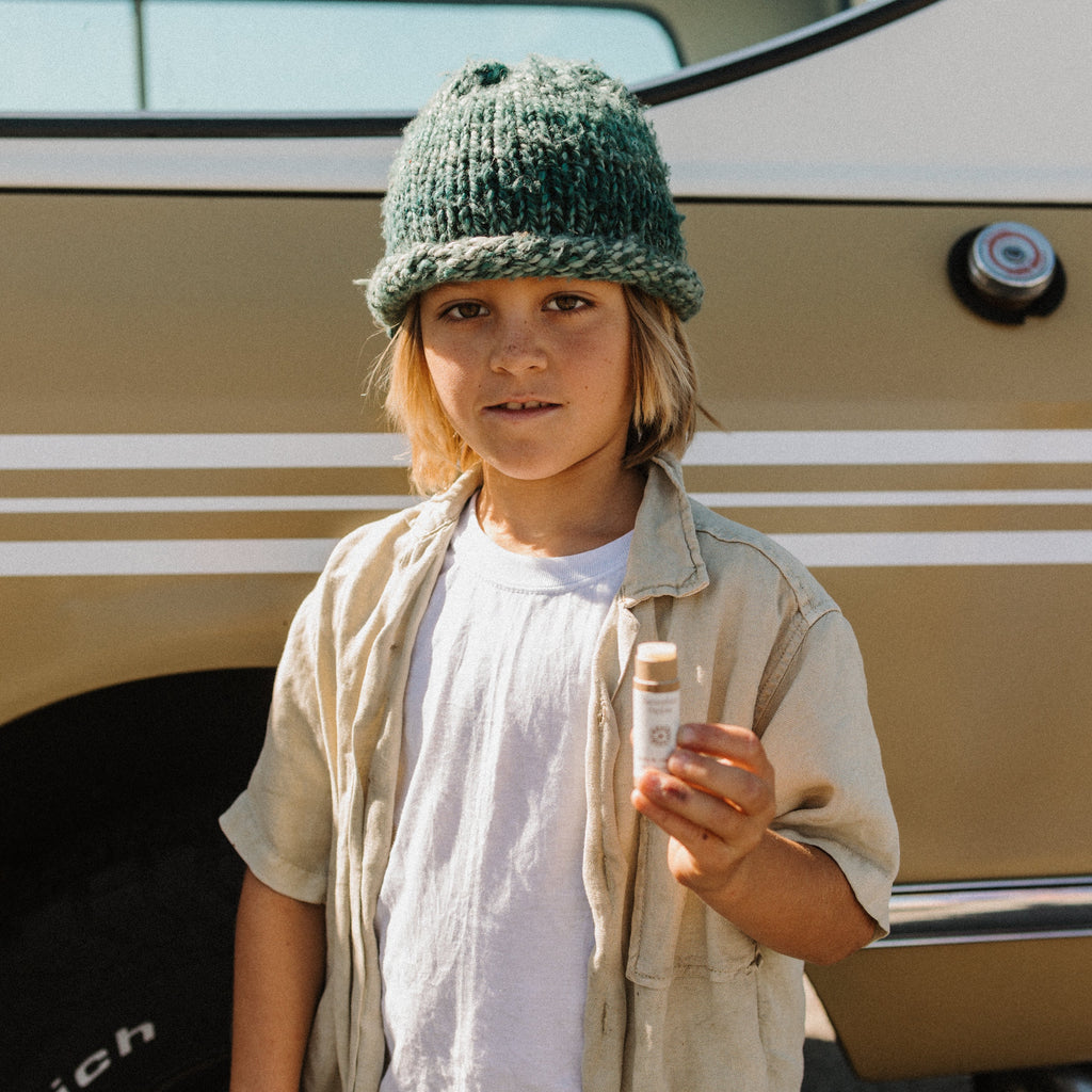 A child in a green knit beanie and light shirt holds a small bottle of Honeybee Hippie vanilla & chamomile tallow lip butter while standing next to a tan vehicle with white stripes.