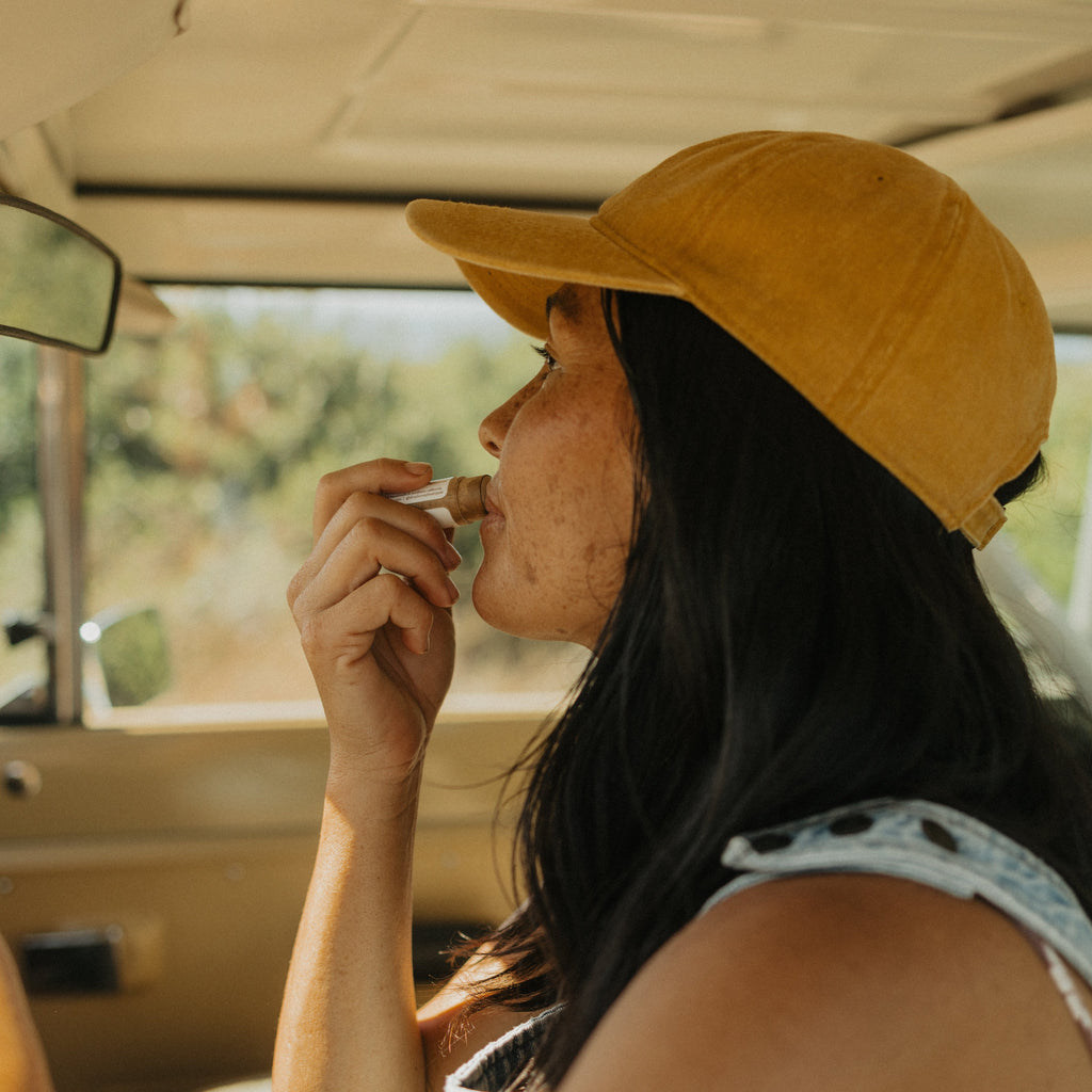 A woman in a yellow cap applies Honeybee Hippie vanilla & chamomile tallow lip butter while sitting in her car, sunlight streaming through the window.