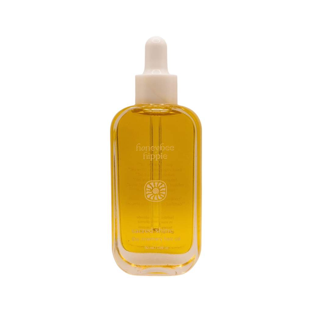 A clear glass bottle of golden yellow hair oil with a white dropper cap; the label reads "Honeybee Hippie" and "sacred shine hair oil" with additional text below.