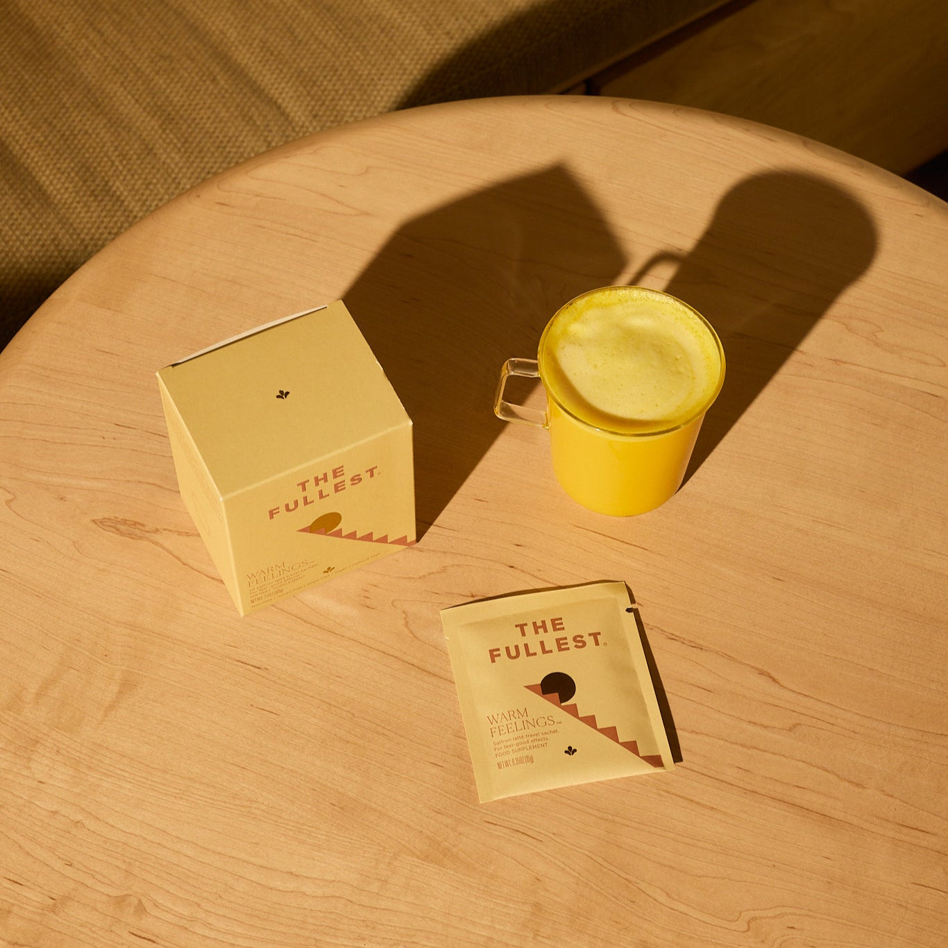 A yellow mug of caffeine-free Saffron Latte sits on a round wooden table beside a beige box and packet labeled "The Fullest Saffron Latte 10 Single-Serving Sachets.