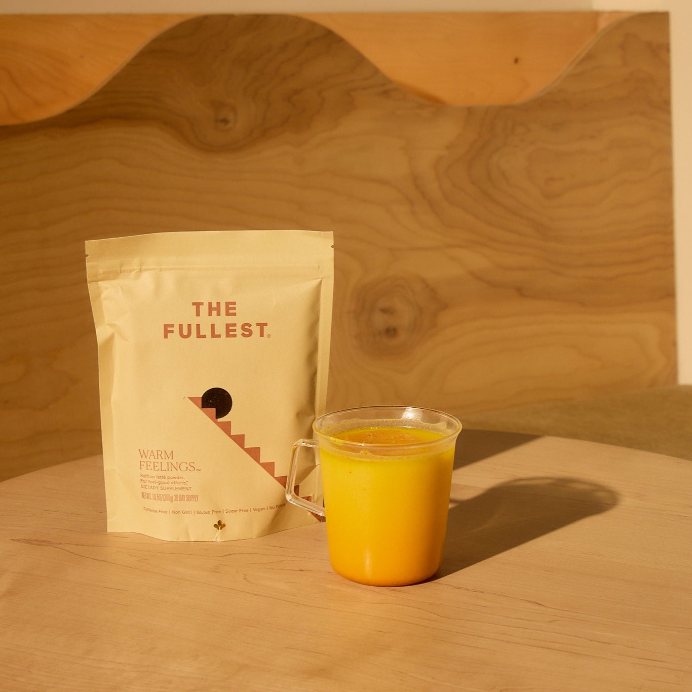 A beige “The Fullest” Saffron Latte 30-Day Supply packet stands on a light wooden table next to a clear mug of yellow, caffeine-free saffron latte, with a wood panel featuring natural grain in the background.