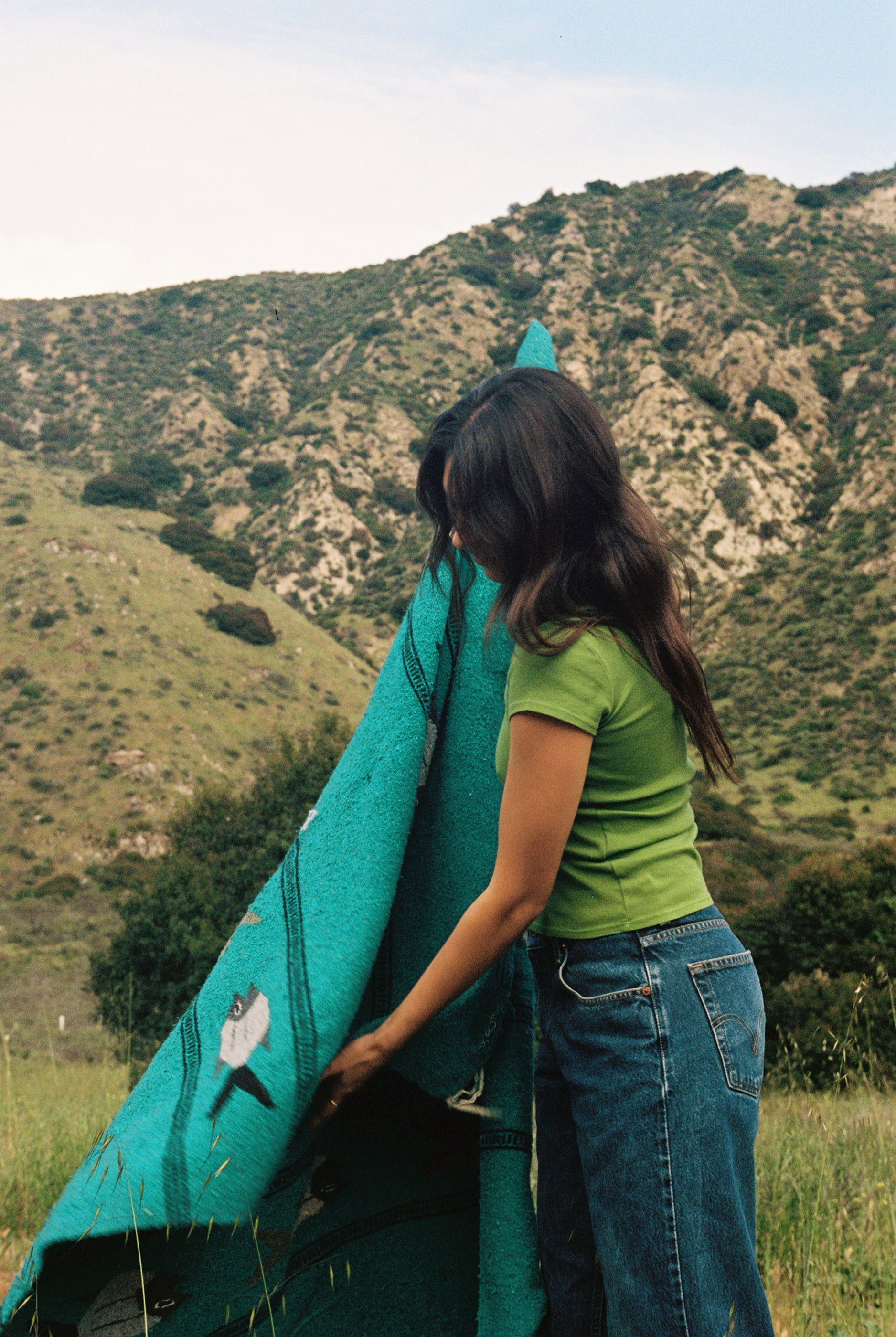 A woman stands in a grassy field by rolling hills, cradling a turquoise blanket. She wears the Bebe Organic Tee by LA Relaxed paired with a denim skirt, her dark hair cascading down. The lush hills and partly cloudy sky provide a serene backdrop to this organic cotton moment.