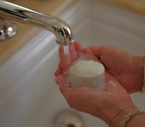 A person holds the bāeo bare bar under running water from a faucet, creating bubbles in their hands over a sink for gentle cleansing.