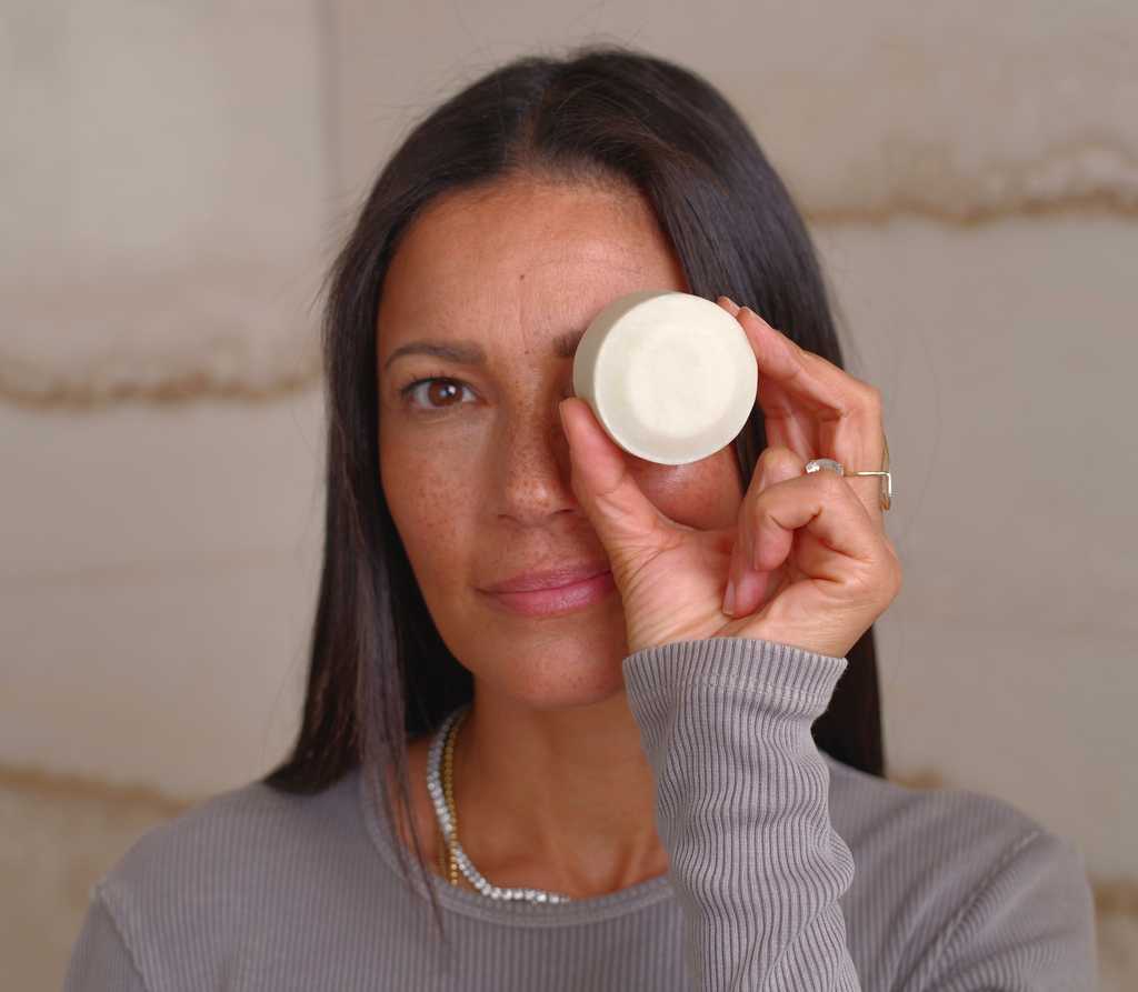 A woman with long dark hair and a gray ribbed top holds a bāeo bare bar—a round white shampoo and body wash bar—up to one eye, looking directly at the camera against a neutral background.