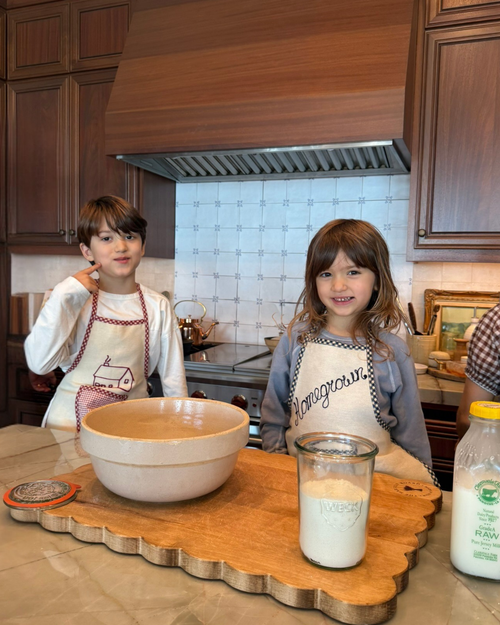 Two kids in Baile x Casa Carini's Baile Homegrown Aprons stand smiling in the kitchen behind a counter with a wooden cutting board, mixing bowl, flour jar, and milk bottle, ready to bake.