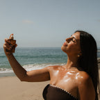 A woman stands on a sandy beach, eyes closed and smiling, as she spritzes Honeybee Hippie's clear skin hydrosol on her face and shoulders. The ocean and clear sky form a serene backdrop.