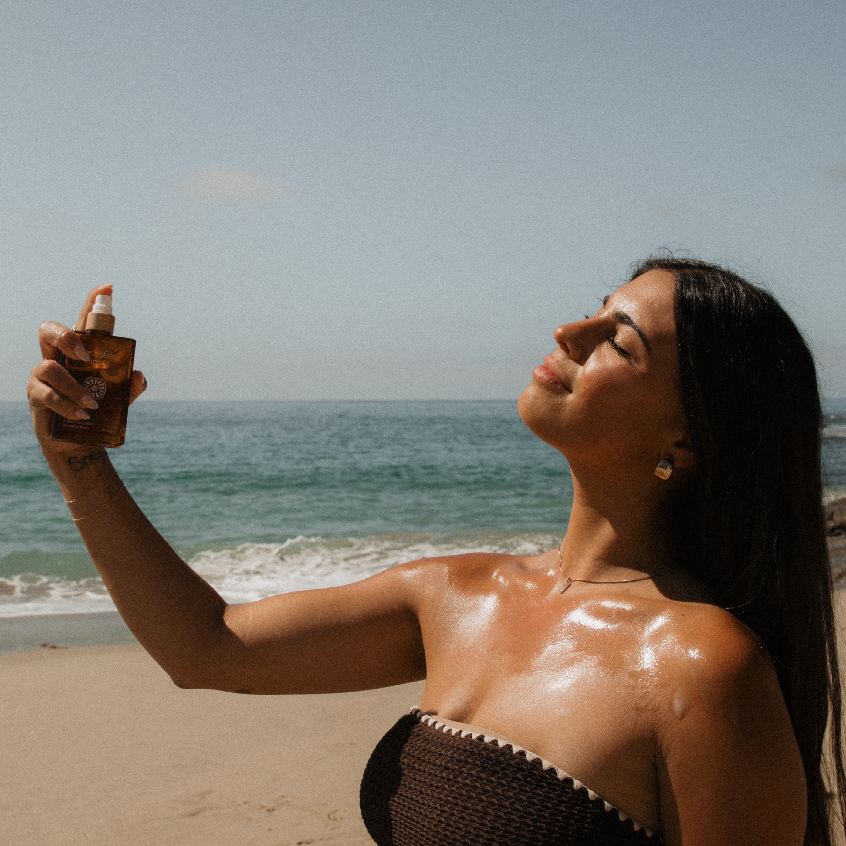 A woman stands on a sandy beach, eyes closed and smiling, as she spritzes Honeybee Hippie's clear skin hydrosol on her face and shoulders. The ocean and clear sky form a serene backdrop.