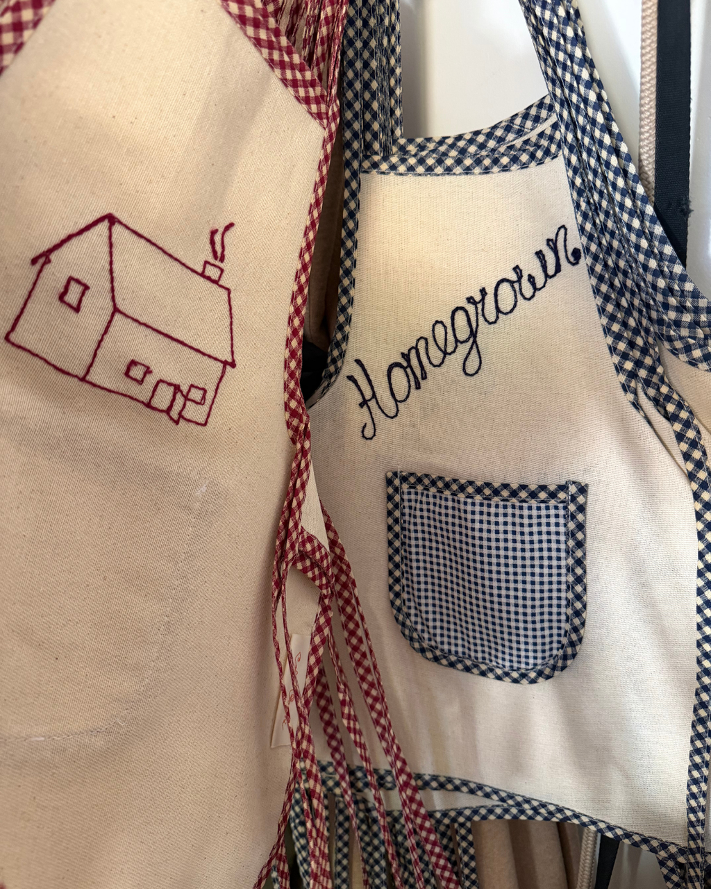 Two Baile x Casa Carini Homegrown Aprons hang side by side: one is handmade cotton with a red embroidered house; the other, a children's apron, features blue gingham trim, a checkered pocket, and "Homegrown" in blue script.