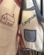 Two Baile x Casa Carini Homegrown Aprons hang side by side: one is handmade cotton with a red embroidered house; the other, a children's apron, features blue gingham trim, a checkered pocket, and "Homegrown" in blue script.
