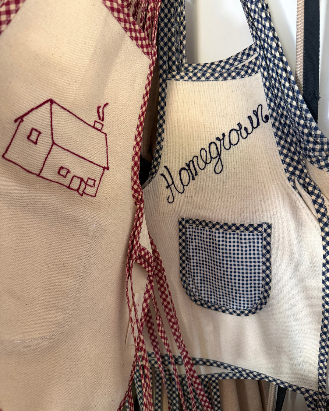 Two Baile x Casa Carini Homegrown Aprons hang side by side: one is handmade cotton with a red embroidered house; the other, a children's apron, features blue gingham trim, a checkered pocket, and "Homegrown" in blue script.