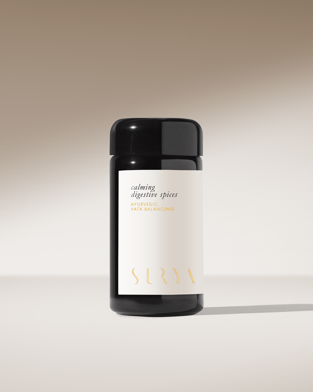A black cylindrical jar labeled "Calming Digestive Spices" by Surya sits on a smooth surface against a beige gradient background—ideal as an Ayurvedic Vata-balancing spice blend for dahl or seasoning vegetables.