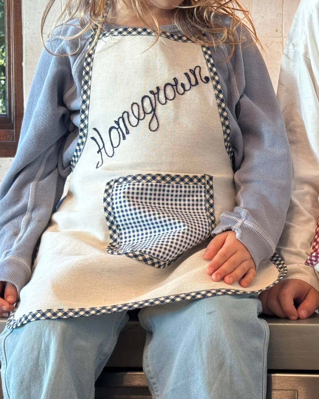 A child in a blue shirt and a Baile x Casa Carini "Homegrown" cotton apron with a checkered pocket sits indoors next to someone in white.