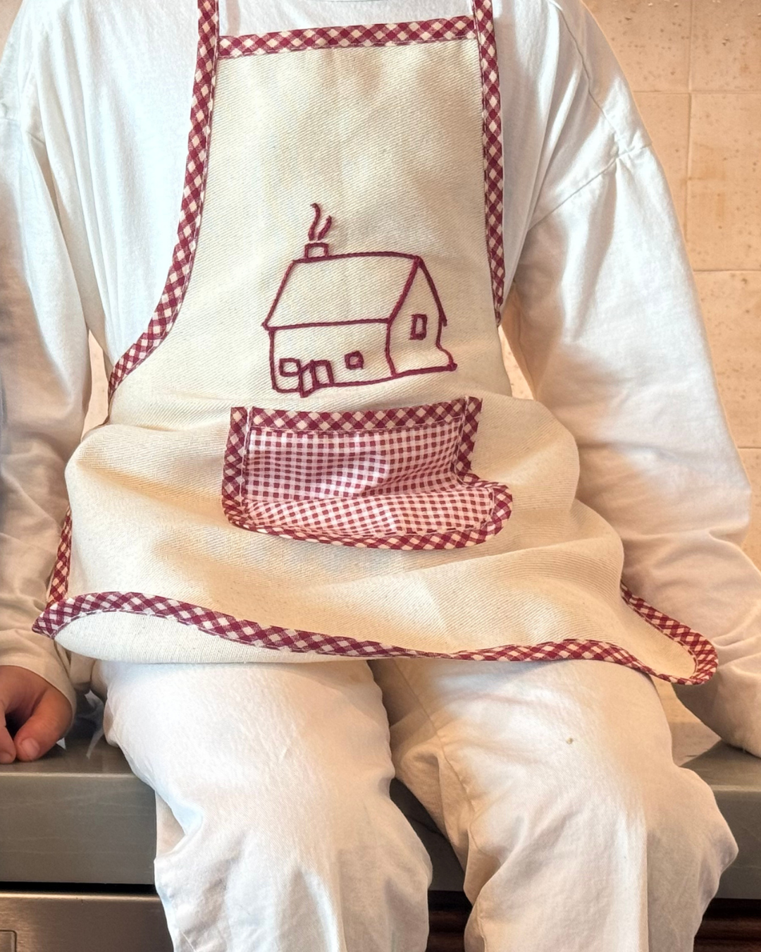 A person in all white sits on a kitchen counter, wearing the cream Baile Homegrown Apron by Baile x Casa Carini—handmade cotton with red checkered trim and a pocket stitched with a house outline.