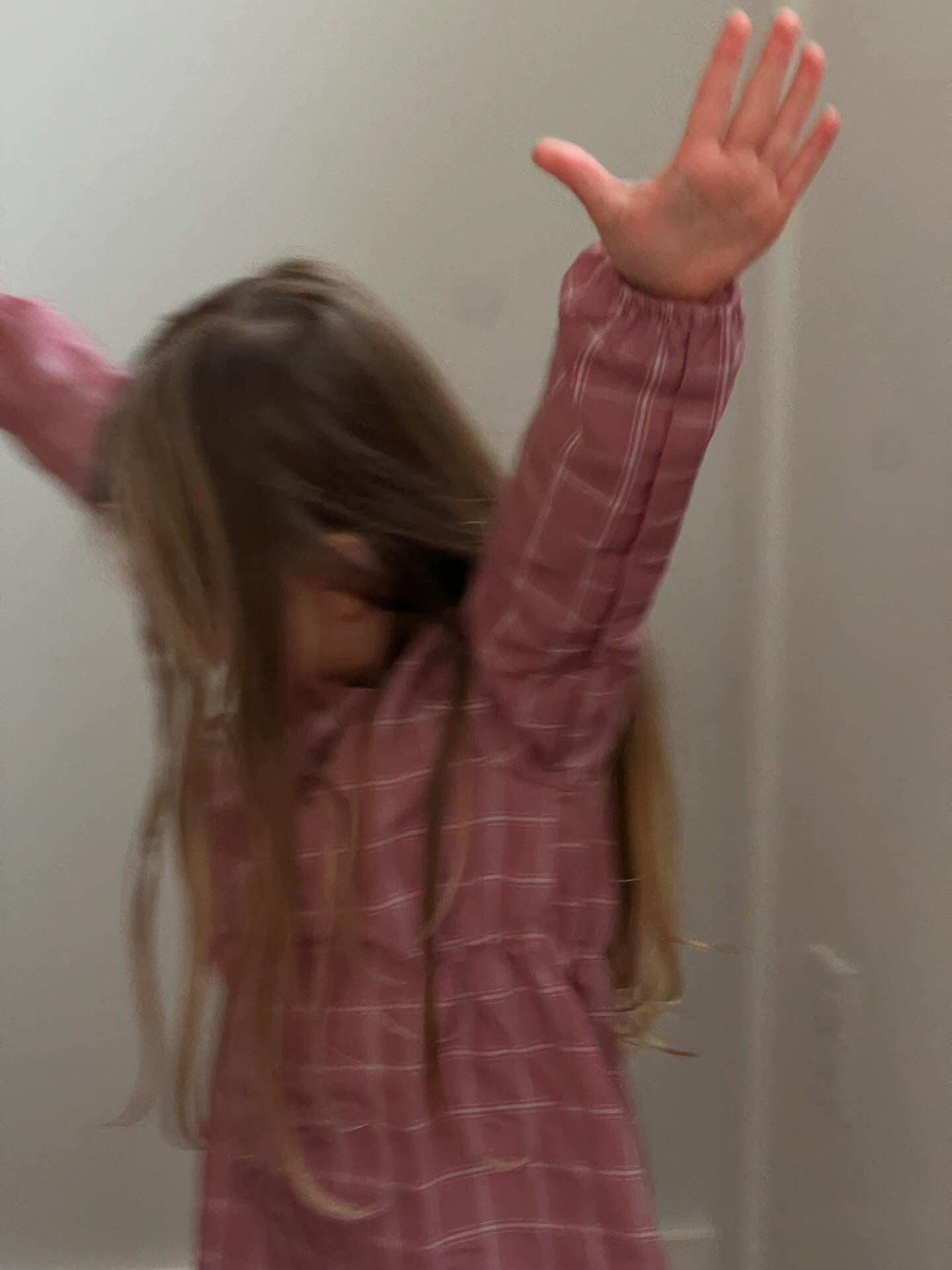 A child wearing the cabane childrenswear's DAY DRESS plaid, handmade in Vancouver, is joyfully raising their arms. Their long hair partially obscures their face, with a simple neutral wall in the background enhancing the playful and energetic atmosphere.