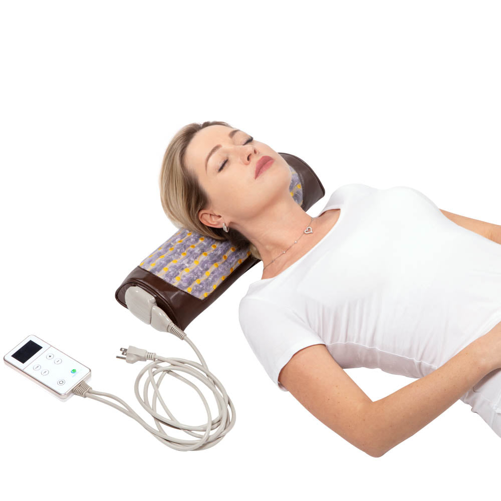 A woman is lying down with her head resting on a HealthyLine Amethyst Bolster Firm | Heated InfraMat Pro®. The device, featuring a patterned brown surface and a control panel attached via a cord, promotes natural healing. She appears relaxed with eyes closed, wearing a white shirt and a delicate necklace.