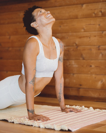 A person with short hair and tattooed arms is performing a yoga pose on the Harda and Madder Root Ayurvedic Yoga Mat from Bennd Yoga, crafted from natural rubber latex. Clad in a white sports tank and pants, they are situated in a wooden-floored room, gazing upwards with a smile that embodies superior performance.