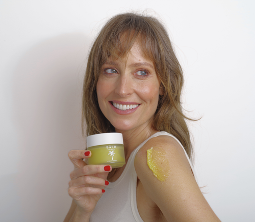 A smiling woman with light brown hair applies bāeo bare butter, an organic hydrating balm for dry or sensitive skin, to her shoulder. She wears a beige tank top and red nail polish, standing against a plain white background.