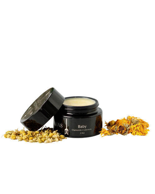 A small dark jar of Wild Mamas Baby- Wild Bison Tallow Balm (2 oz) with the lid resting nearby, surrounded by dried chamomile and calendula—gentle care for newborn skin on a white background.