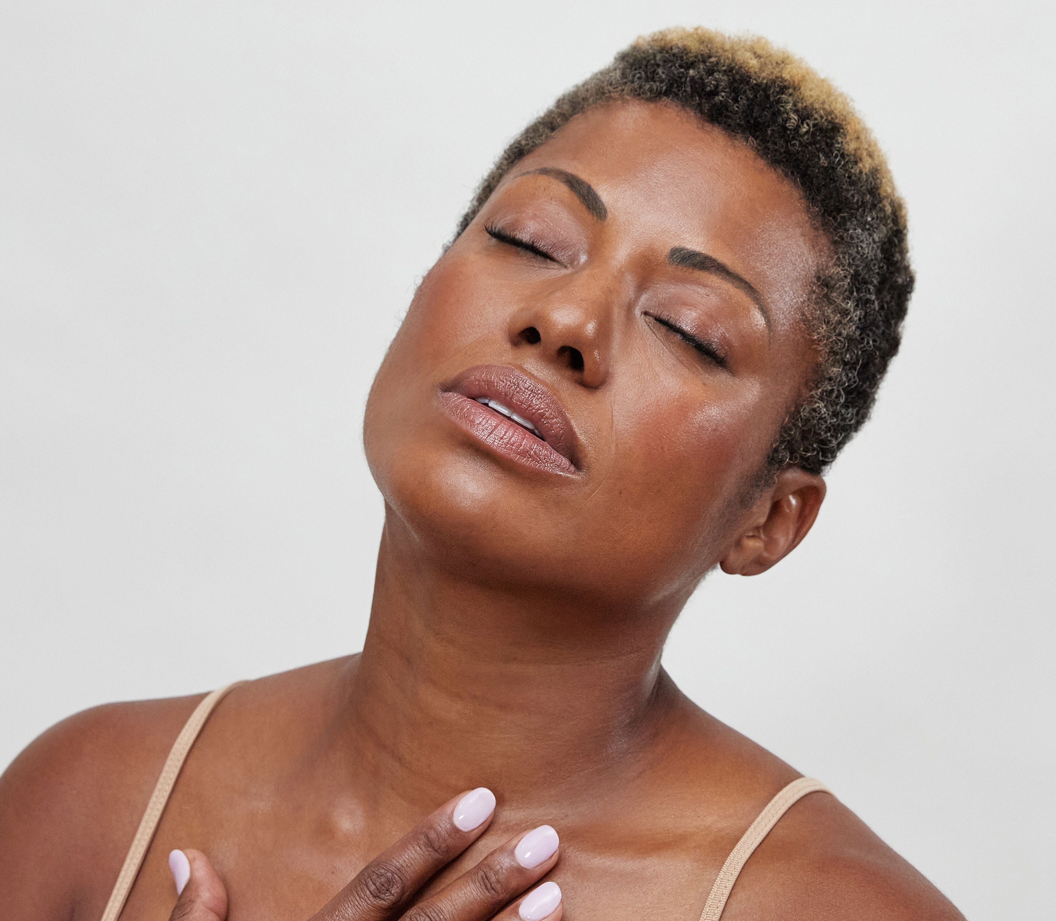 A woman with short hair and closed eyes tilts her head back, her skin glowing with a dewy finish from bāeo's bare tint 'palisades', radiating calm and relaxation against a plain light background.