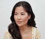 A woman with long dark hair, in a gold necklace and white-yellow patterned top, looks to the side against a light background, her lips and cheeks glowing with bāeo's bare tint 'palisades' for a natural dewy finish.
