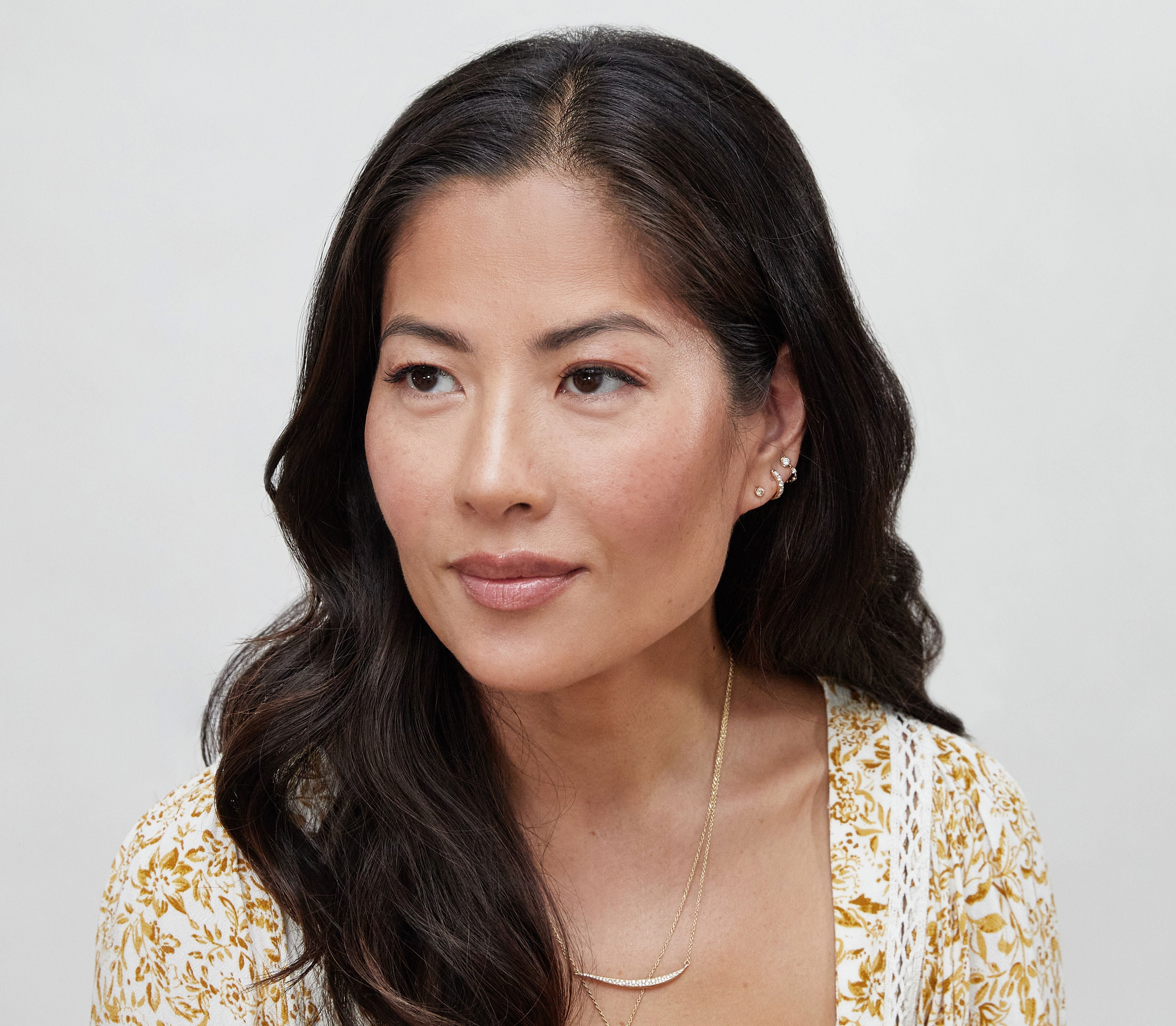 A woman with long dark hair, in a gold necklace and white-yellow patterned top, looks to the side against a light background, her lips and cheeks glowing with bāeo's bare tint 'palisades' for a natural dewy finish.