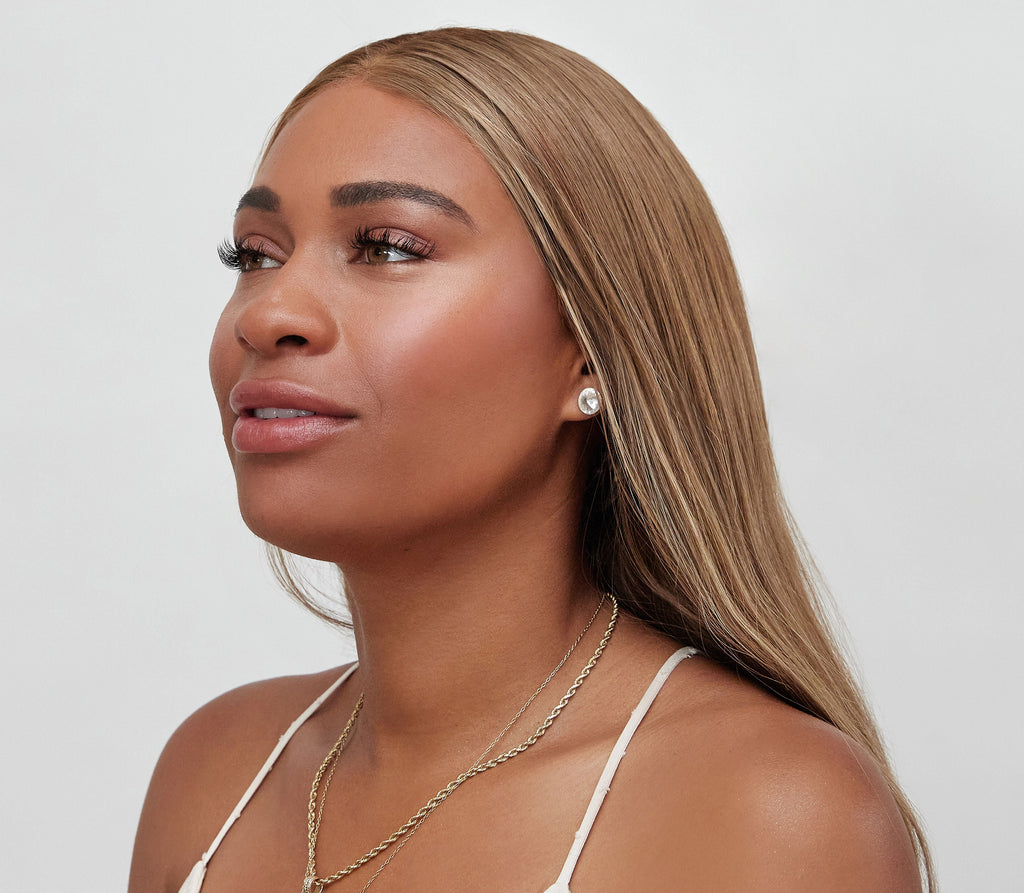 A woman with long, straight, light brown hair looks off to the side, wearing stud earrings and a gold chain necklace. She wears bāeo's bare tint 'palisades' for a natural dewy look that stands out against her white top and the light background.