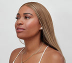 A woman with long, straight, light brown hair looks off to the side, wearing stud earrings and a gold chain necklace. She wears bāeo's bare tint 'palisades' for a natural dewy look that stands out against her white top and the light background.