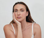 A woman with long brown hair rests her face on her hand, wearing bāeo's bare tint 'palisades' for a natural dewy finish on lips and cheeks. She gazes at the camera in a white top, gold necklace, and small hoop earrings against a plain background.