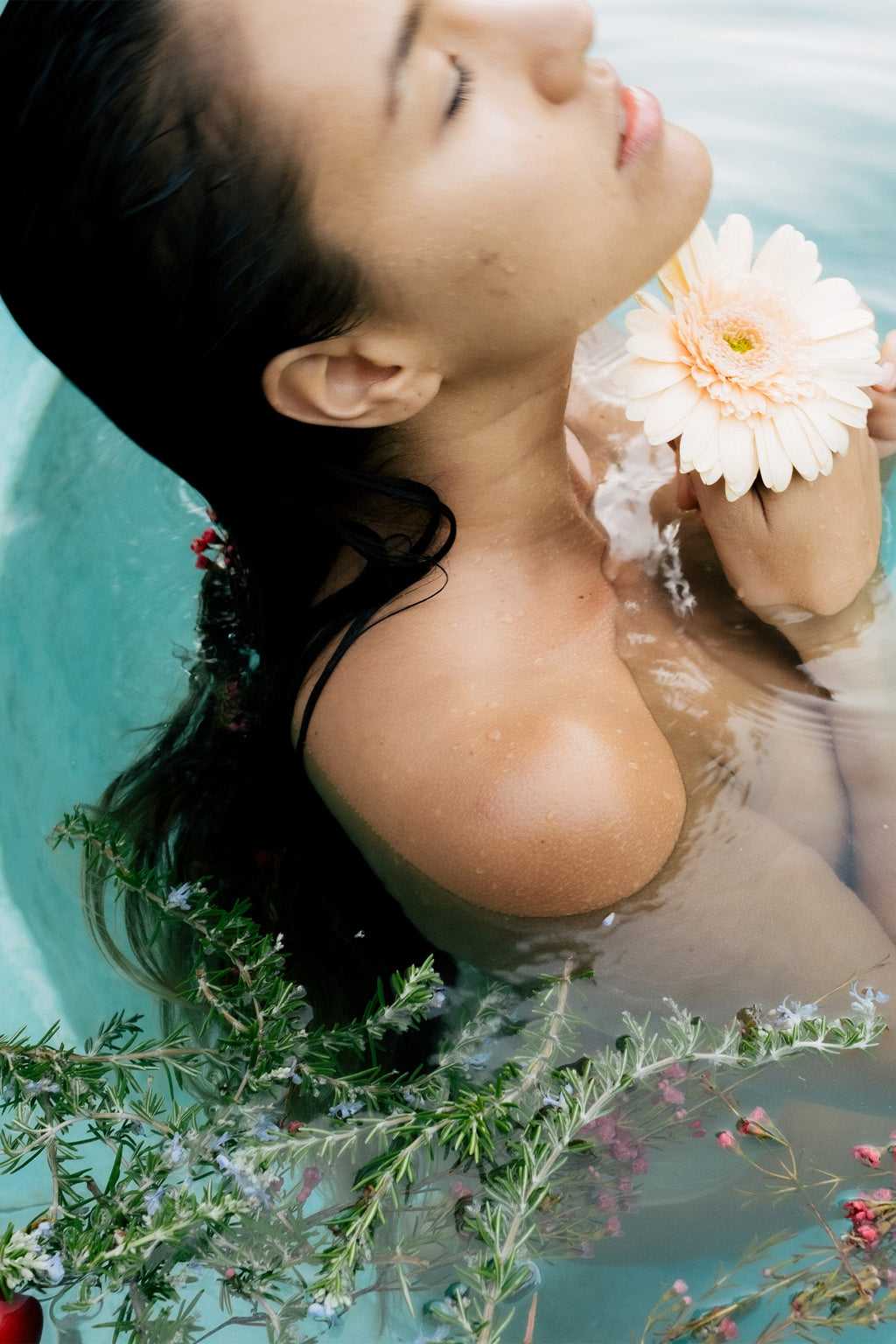 A woman with wet hair relaxes in a Surya Cooling Bath Soak, eyes closed in calm reflection, holding a pale peach gerbera daisy at her collarbone as green foliage and small flowers float around her, harmonizing emotions.