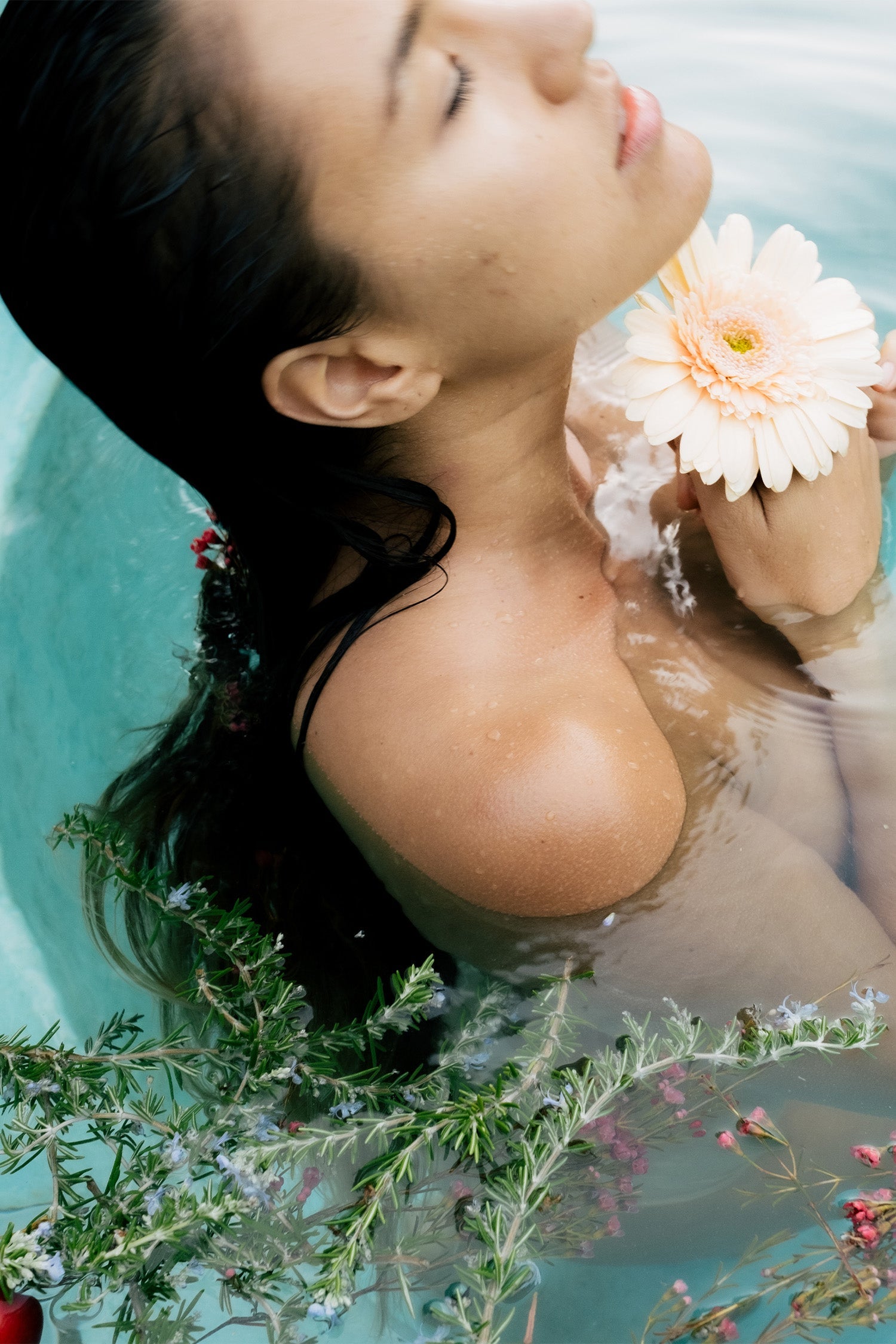 A woman with wet hair relaxes in a Surya Cooling Bath Soak, eyes closed in calm reflection, holding a pale peach gerbera daisy at her collarbone as green foliage and small flowers float around her, harmonizing emotions.